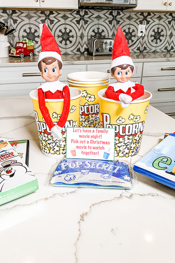 Two elf on a shelf dolls in popcorn cups with a note in front of them that says Let's have a family movie night! Pick out a Christmas movie to watch together. Next to the note are stacks of Christmas movies and a bag of unpopped popcorn. 