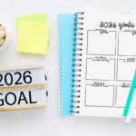 2026 goals worksheet in a spiral notebook on a white desk with teal and white pens, a succulent plant, sticky notes, and a “2026 Goal” marquee sign beside it.