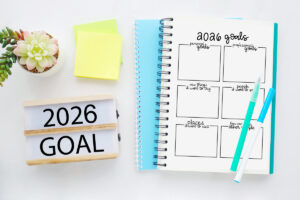 2026 goals worksheet in a spiral notebook on a white desk with teal and white pens, a succulent plant, sticky notes, and a &ldquo;2026 Goal&rdquo; marquee sign beside it.