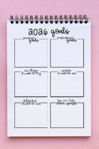 2026 goals worksheet in a spiral notebook on a pink background, showing blank boxes for personal goals, professional goals, new things to try, people to see, places to visit, and ways to help others.