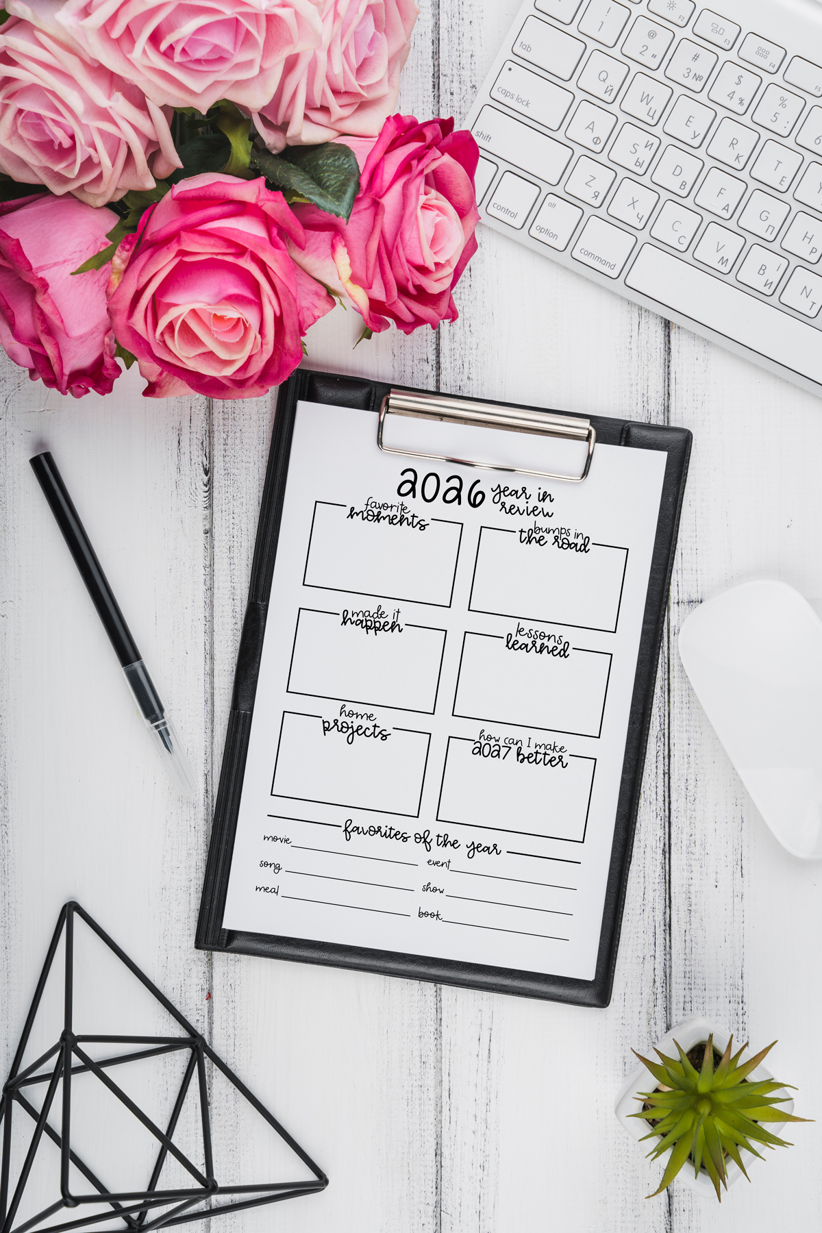 2026 year-in-review worksheet on a black clipboard styled on a white wooden desk with pink roses, a keyboard, a pen, geometric decor, and a small succulent.