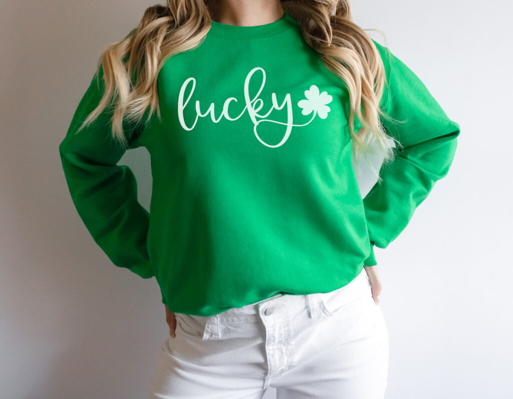 This image represents one of the free lucky SVG files you can download at the end of the post. It shows a girl from the neck down to her legs wearing a green shirt and white jeans. Her green shirt shows the cursive version of the free SVG you can download at the end - the design is the word lucky in cursive with a green shamrock coming off the tail of the y. The text is in white.