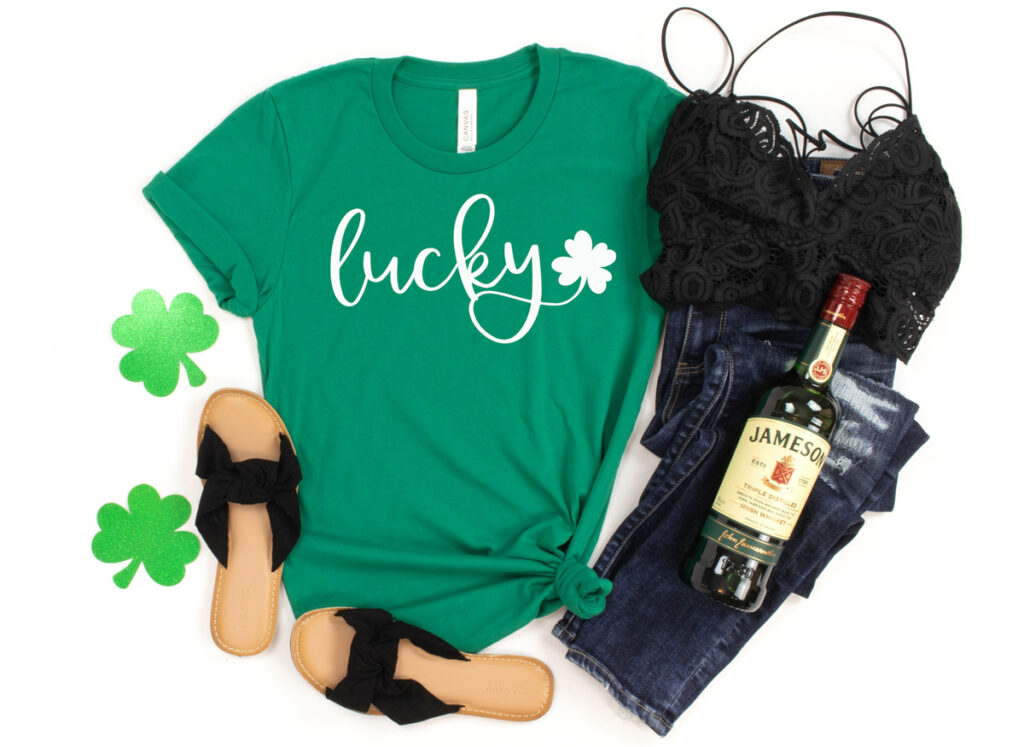 This shirt shows one of the free lucky svg that can be downloaded at the end of this blog post. It has a green shirt with the word lucky in white cursive with a small shamrock. There are also a pair of sandals, jeans, a black tank top, two glittery paper shamrocks, and a bottle of Jameson.