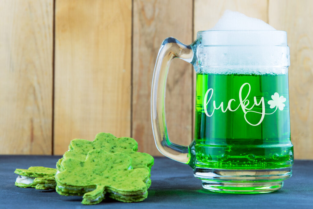 This image shows one of the two lucky svg files you can download. It is shown on a beer stein with green beer in it, sitting next to a shamrock shaped sandwich. The word lucky is etched in cursive on the beer stein with a shamrock coming off the tail of the y in lucky.