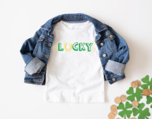 This image shows one of the free lucky SVG files you can download at the end of this blog post. This version is the print lucky with a horseshoe replacing the letter u. The horseshoe is yellow and facing up like a U. The text for the rest of lucky is in green. In this image, it is on a white shirt with a jean jacket over top. In the bottom right corner are some gold coins and some glittery paper shamrocks.
