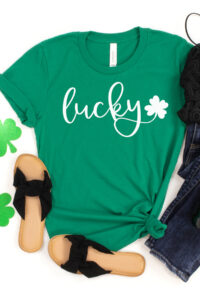 This shirt shows one of the free lucky svg that can be downloaded at the end of this blog post. It has a green shirt with the word lucky in white cursive with a small shamrock. There are also a pair of sandals, jeans, a black tank top, two glittery paper shamrocks, and a bottle of Jameson.