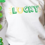 This image shows one of the free lucky SVG files you can download at the end of this blog post. This version is the print lucky with a horseshoe replacing the letter u. The horseshoe is yellow and facing up like a U. The text for the rest of lucky is in green. In this image, it is on a white shirt on a girl (showing a it zoomed in only from her neck to her legs). In her hand she is holding a St. Patrick’s Day themed glittery paper garland of a green hat and shamrocks.