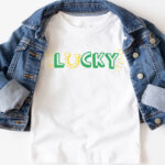 This image shows one of the free lucky SVG files you can download at the end of this blog post. This version is the print lucky with a horseshoe replacing the letter u. The horseshoe is yellow and facing up like a U. The text for the rest of lucky is in green. In this image, it is on a white shirt with a jean jacket over top. In the bottom right corner are some gold coins and some glittery paper shamrocks.