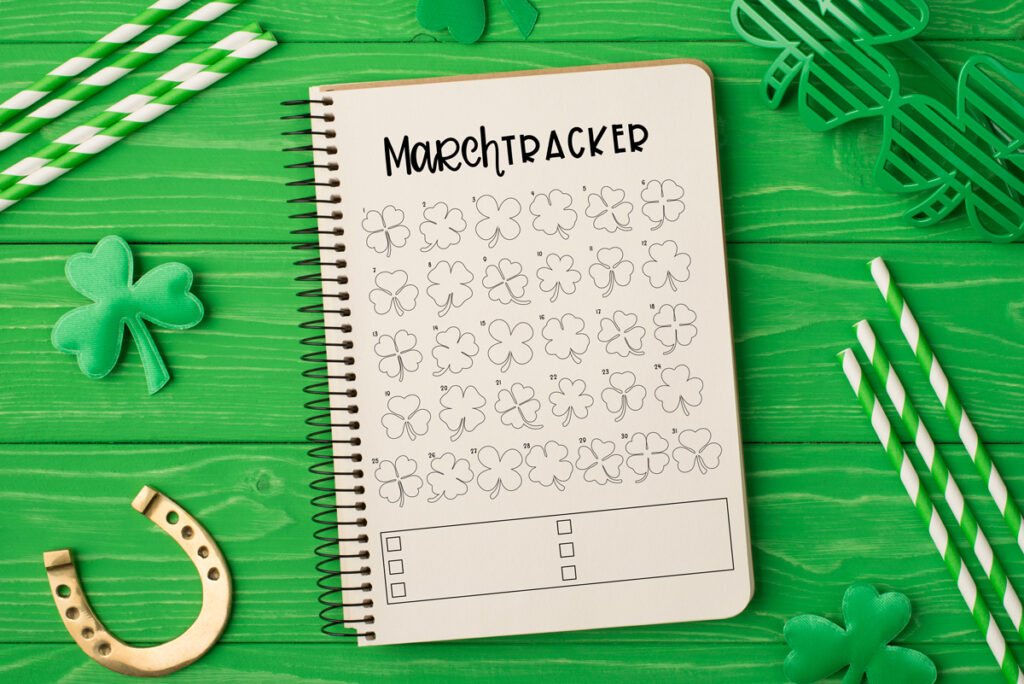 This image shows the March mood tracker available to download at the end of this post. It is is an image of the March mood and habit tracker on a notebook on green stained wood with St. Patrick’s day things around it including a green felt shamrock, a gold horseshoe, green and white stripped straws, and green clover shaped glasses.