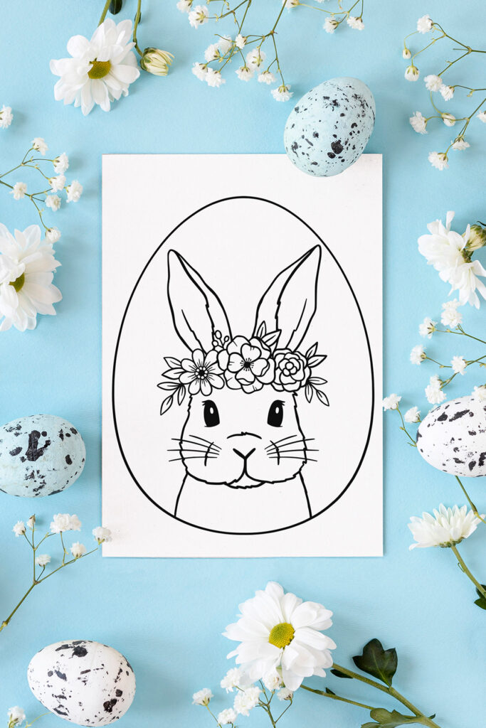 This image shows one of the Easter Egg coloring pages designs you can download for free at the end of this blog post. This image shows a cute bunny with a floral headband in an easter egg on a coloring page. It is on a blue background surrounded by flowers and Easter eggs.