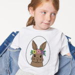 This is an image of a little girl wearing one of the free 60 Easter Egg SVGs you can download at the end of this blog post. The girl has a white shirt on with a white egg with a bunny wearing a floral crown in the middle of the egg.