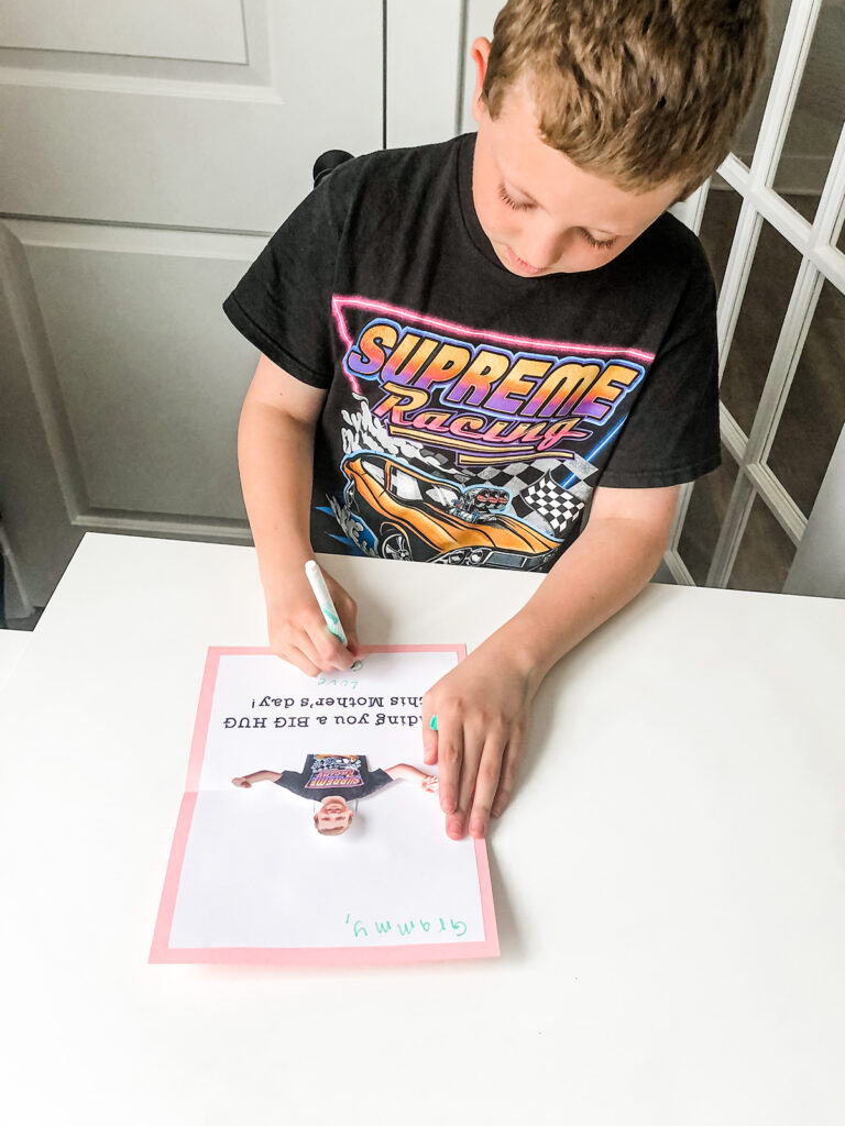 This image shows a child adding their greeting and signature to the inside of the pop up card. You see what the inside of the pop up card looks like. It has the text, “sending you a BIG HUG this Mother’s day!”