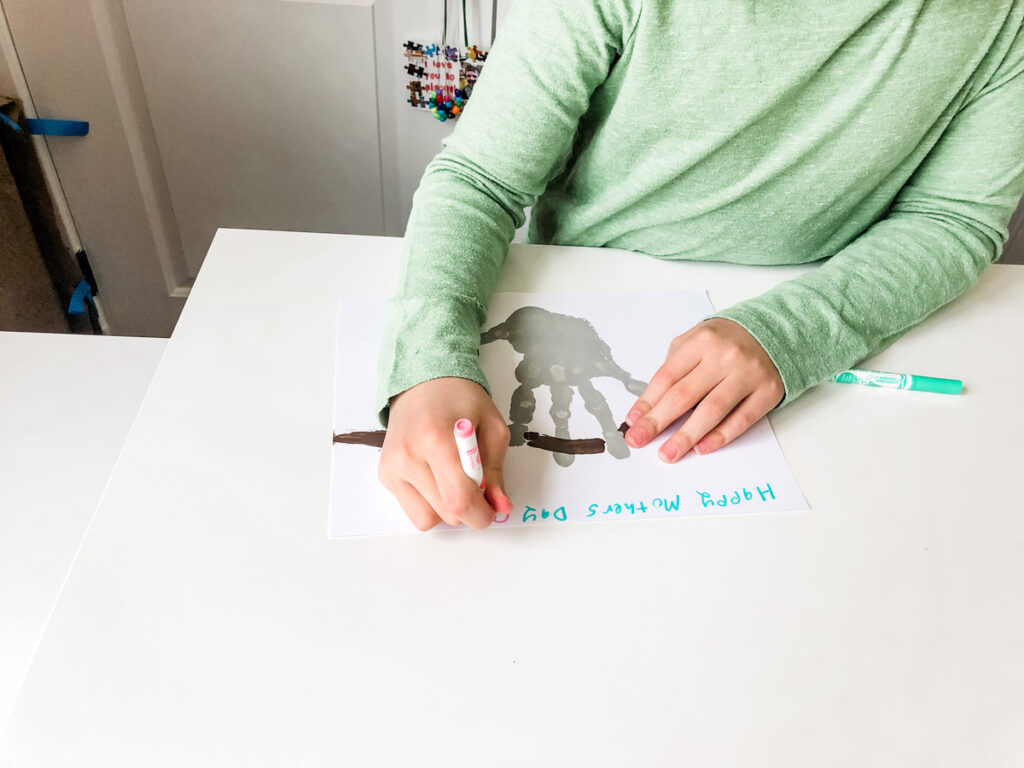 This image shows a child writing Happy Mother's Day at the top of the sloth handprint craft.