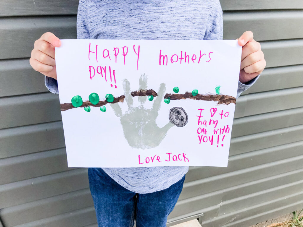 This image shows a child holding up the completed sloth handprint craft for Mother's Day. At the top of the craft he text says Happy Mother's Day. Below that it says I love hanging out with you. Love, Jack. Next to the text is a gray handprint shape that has been turned into a sloth with a sloth head made from the free sloth head template. The sloth is hanging from a hand drawn brown branch with green fingertip leaves.