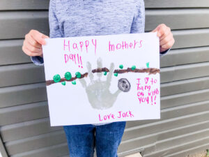 This image shows a child holding up the completed sloth handprint craft for Mother's Day. At the top of the craft he text says Happy Mother's Day. Below that it says I love hanging out with you. Love, Jack. Next to the text is a gray handprint shape that has been turned into a sloth with a sloth head made from the free sloth head template. The sloth is hanging from a hand drawn brown branch with green fingertip leaves.