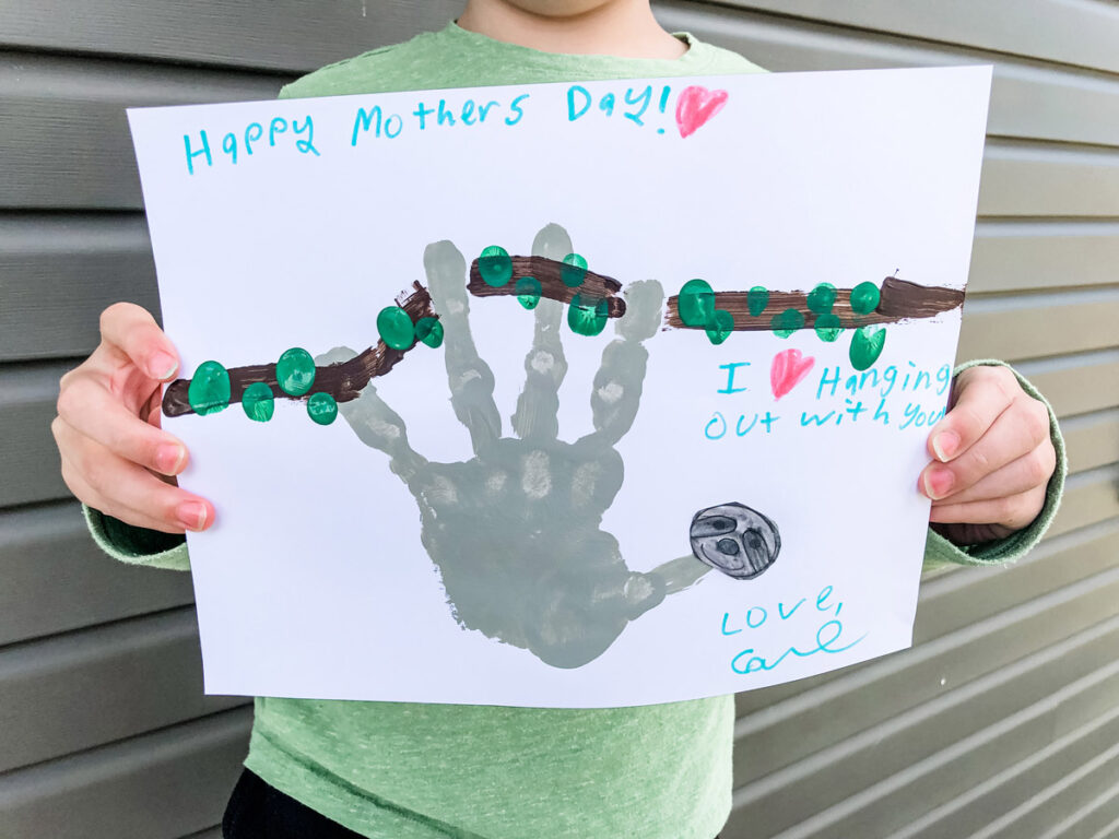 This image shows a child holding up the completed sloth handprint craft for Mother's Day. At the top of the craft he text says Happy Mother's Day. Below that it says I love hanging out with you. Love, Carl. Next to the text is a gray handprint shape that has been turned into a sloth with a sloth head made from the free sloth head template. The sloth is hanging from a hand drawn brown branch with green fingertip leaves.