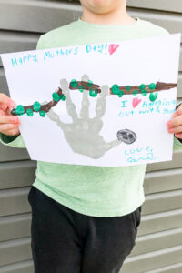 This image shows a child holding up the completed sloth handprint craft for Mother's Day. At the top of the craft he text says Happy Mother's Day. Below that it says I love hanging out with you. Love, Carl. Next to the text is a gray handprint shape that has been turned into a sloth with a sloth head made from the free sloth head template. The sloth is hanging from a hand drawn brown branch with green fingertip leaves.