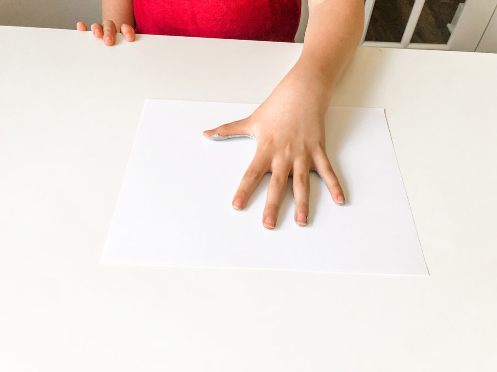 In this image, a child is placing their paint colored hand in the center of a blank piece of white paper.