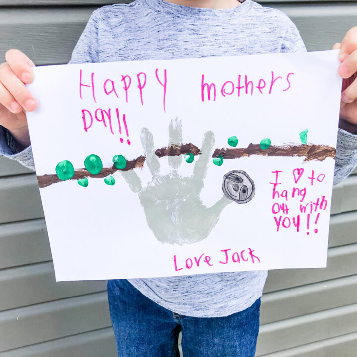 This image shows an example of the completed sloth handprint Mother's Day craft. This image is to show the final product to go with the directions. A child is holding the craft. At the top of the craft, it says Happy Mother's Day!! Below that is the image of a sloth created with a handprint. Next to that and below it says I love (heart image) to hang out with you. Love, Jack.