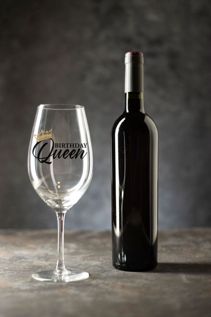 This image shows one of the free birthday SVG files you can get in this blog post. It shows a wine bottle sitting next to a wine glass. On the wine glass, it has the birthday queen SVG.