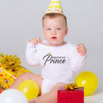This image shows an example with one of the free svgs you can get in this blog post. It shows a little boy wearing a white onesie with one of the free svgs, Birthday Prince. He is wearing a birthday hat and surrounded by balloons and presents.
