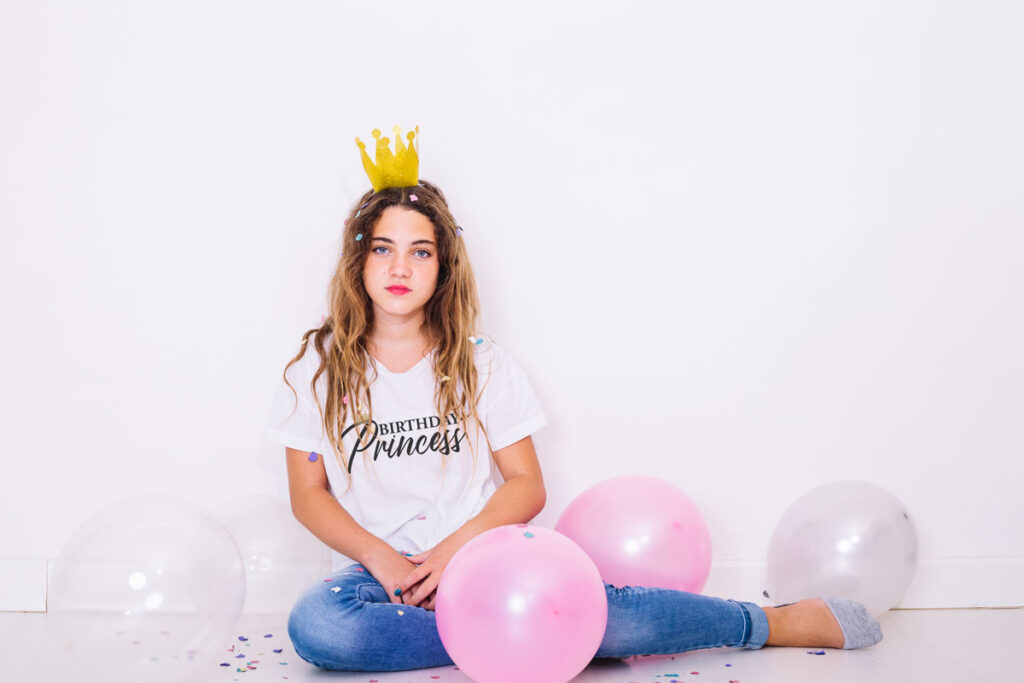 This image shows an example with one of the free svgs you can get in this blog post. It shows a girl wearing a white t-shirt with one of the free svgs, Birthday Princess. She is wearing a crown and is surrounded by pink and white balloons.