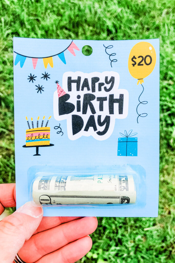 This image shows the free chapstick money holder template holding a $20 bill. The template says Happy Birthday with a balloon in the top right corner with $20. The template also includes a birthday cake with candles, a gift box, a banner, and a party hat on the B.