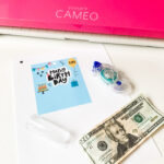 This image shows the materials required to make the chapstick money holder template. It has the printed template, a Craft cutting machine (a Silhouette Cameo), an adhesive, plastic chapstick bubble, and a $20 bill (you can include the amount of money you’d like).