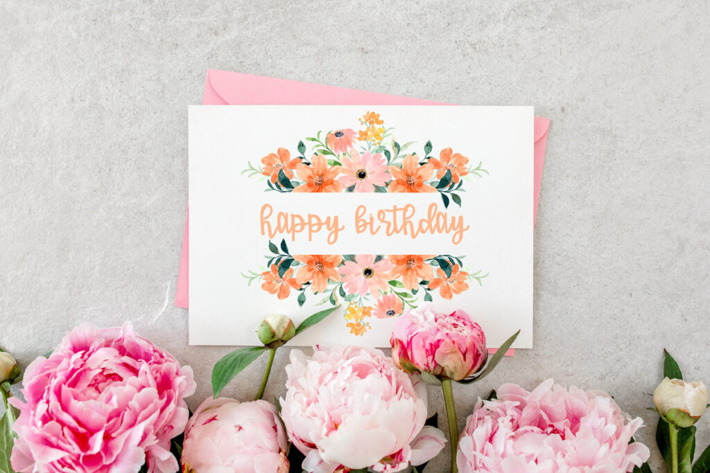 This image shows an example of a flower birthday card you can download for free in this set of 8 free printable birthday cards. This image shows a card with peach, pink, and orange flowers at the top and bottom of a white rectangle with a peach Happy Birthday in the middle of the white rectangle. At the bottom center of the printed card are pretty pink real flowers.