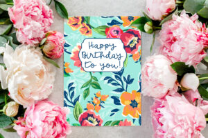 This image shows an example of a flower birthday card you can download for free in this set of 8 free printable birthday cards. This image shows a card with a teal blue background and bright orange, and deep pink flowers with dark blue leaves. In the top middle, it has a white label with a blue Happy Birthday to You! Message. The left and right side of the printed card are pretty pink and white real flowers.