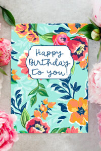 This image shows an example of a flower birthday card you can download for free in this set of 8 free printable birthday cards. This image shows a card with a teal blue background and bright orange, and deep pink flowers with dark blue leaves. In the top middle, it has a white label with a blue Happy Birthday to You! Message. The left and right side of the printed card are pretty pink and white real flowers.