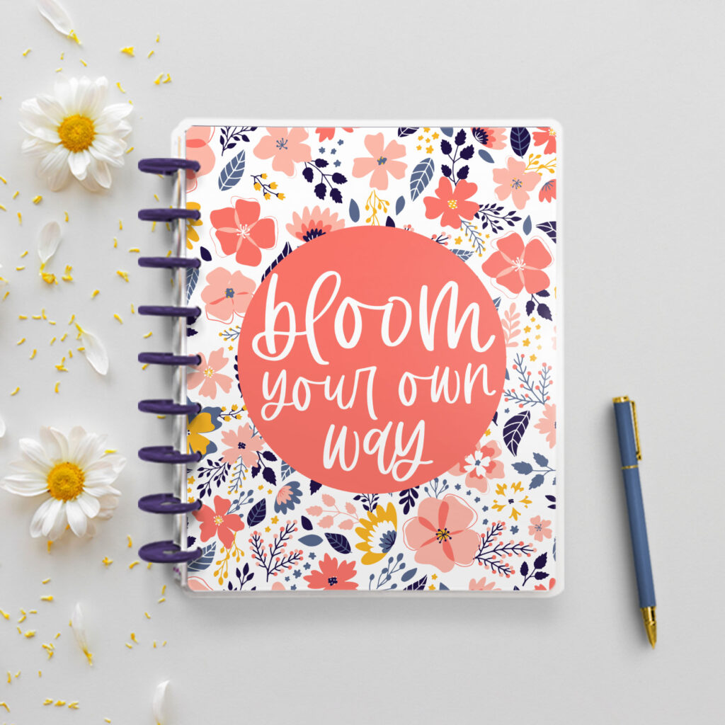 This is an image of a free floral planner cover printable you can download at the end of this blog post. The picture is of a closed disc planner with a floral design on the cover. In the center of the cover, it has the quote, “bloom your own way” on it. To the left are some white daisies and to the right is a blue pen.