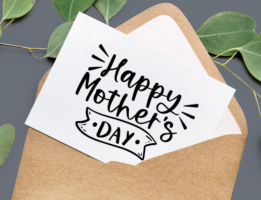 This is an image of the free Happy Mother's Day SVG available to download at the end of this post. The file is shown on a white card sitting in a brown Kraft color envelope with some green leaves behind it. The card itself says Happy Mother's Day.