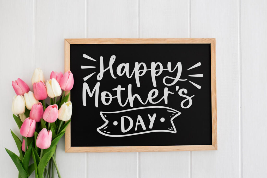 The image is of a chalkboard with the words, Happy Mother's Day written on it. To the left of the chalkboard are some pink and white tulips.