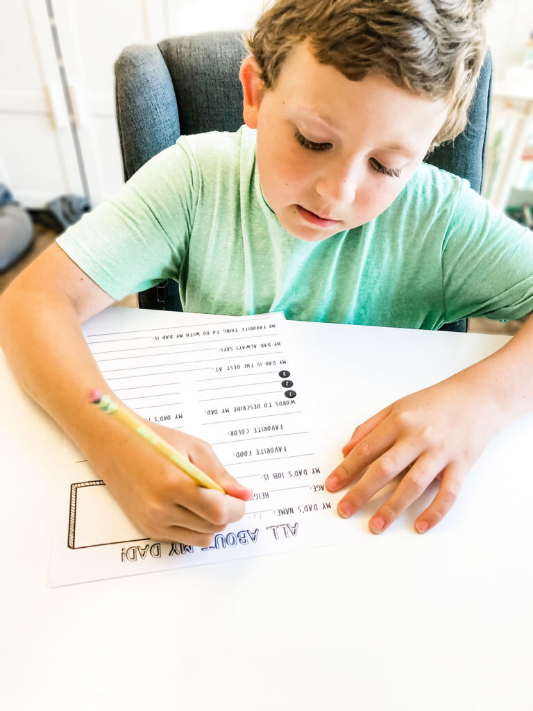 This is an image of a child filling out the Father’s Day questionnaire you download for free at the end of this blog post. This image shows a young boy in a green shirt using a pencil to write in the All About My Dad printable.