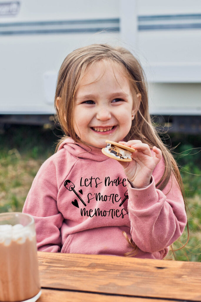 This image shows a little girl eating s'mores at a picnic table. Her shirt has a copy of the making smore memories SVG.