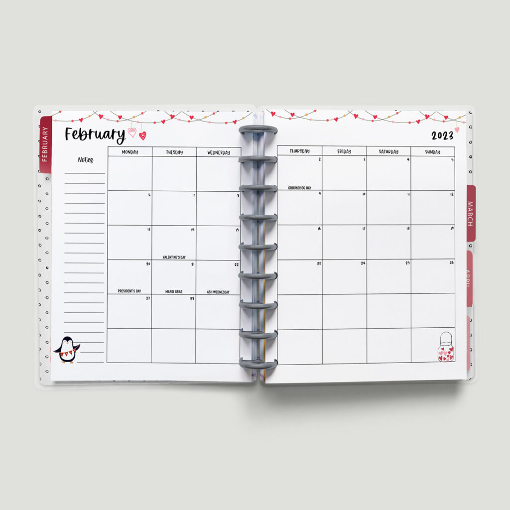 This image is of a planner open to a 2023 calendar with holidays. This is one of the options you can download at the end of this blog post. This image shows a 2023 February calendar with a Monday start. It has a design with hearts, a penguin, and more.