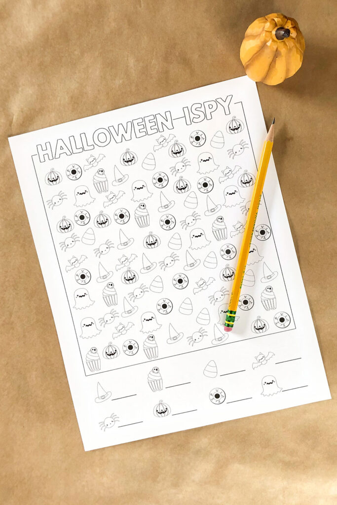 This is an image of the free Halloween I spy in the black and white harder design. It is laying on brown Kraft paper with a small fake orange pumpkin in the top corner and a pencil laying on top of the worksheet. You can download this and three other free Halloween i Spy worksheet options.