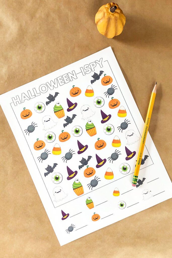 This is an image of the free Halloween I spy in the color, easier design. It is laying on brown Kraft paper with a small fake orange pumpkin in the top corner and a pencil laying on top of the worksheet. You can download this and three other free Halloween i Spy worksheet options.