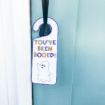 This image shows the you’ve been booed door hanger you can get for free in the You’ve been Booed printable set at the end of this blog post. It’s a white door hanger with a purple outline. It has a little ghost and says, “You’ve been booed!” It is hanging on a front door.