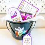 This image shows the You’ve Been Booed printable set being used with a boo basket. It is a picture of a black bucket that says BOO on it. It is filled with Halloween trinkets and treats and has a little gift tag that says “You’ve Been Booed.” In the basket it also has a We’ve Been Booed hanger and you’ve been booed instruction sheet.