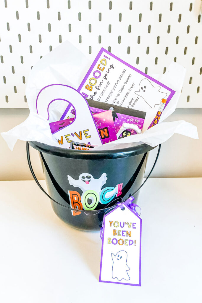 This image shows the You’ve Been Booed printable set being used with a boo basket. It is a picture of a black bucket that says BOO on it. It is filled with Halloween trinkets and treats and has a little gift tag that says “You’ve Been Booed.” In the basket it also has a We’ve Been Booed hanger and you’ve been booed instruction sheet.