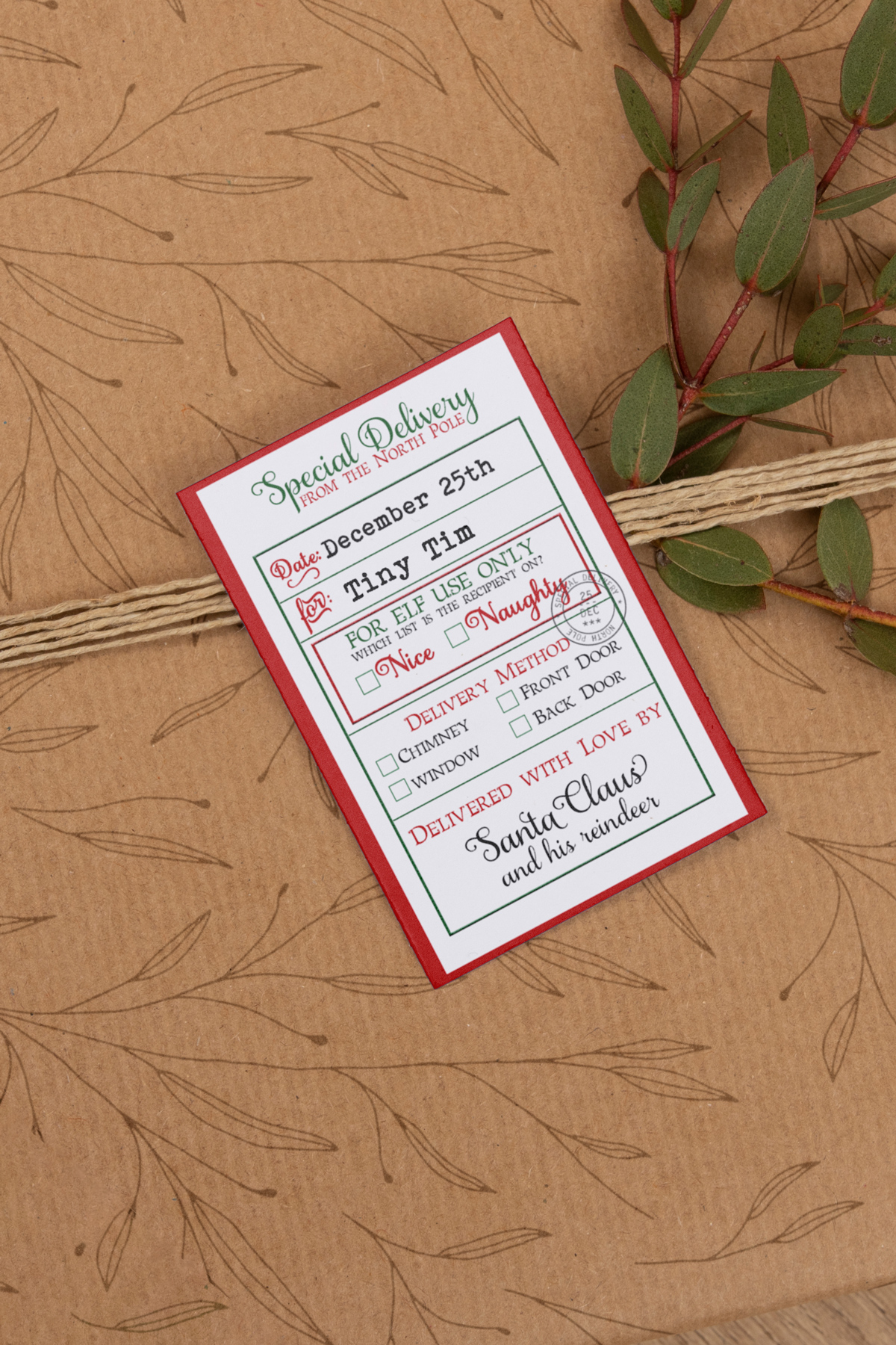 The image shows one of the tags from the Christmas tags from Santa printable set. The tag is being used like a sticker (or label) on a package wrapped with a brown wrapping paper with some brown flowers on it.