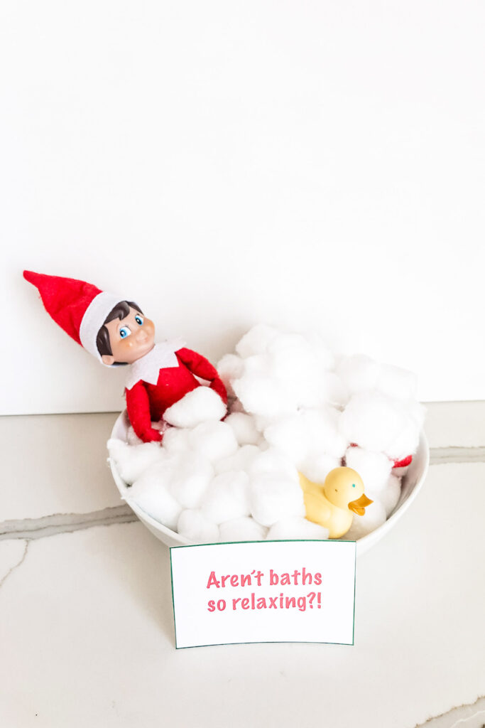 This image shows an Elf on the Shelf taking a fake bath in a bowl with cotton balls and a rubber ducky. There is a sign that says aren’t baths so relaxing?! This is one of the many elf on the shelf ideas for home included in this post.