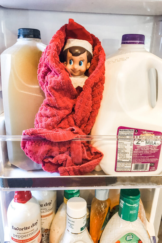 This image shows an Elf on the Shelf dollar stuck in the fridge with a towel wrapped around him like a blanket. It’s one of many easy Elf on the Shelf ideas you can recreate at home.