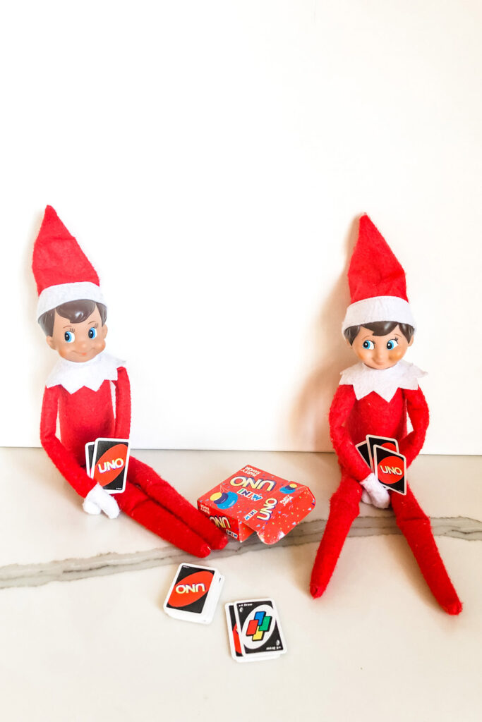 This image shows 2 elf on the shelf dolls playing a game of uno. The cards are miniature in size. This is one of the many elf on the shelf ideas for home included in this post.