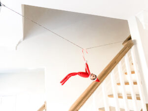 This image shows an elf on the shelf doll hanging from a candy cane across a rope like a zipline. This is one of the many elf on the shelf ideas for home included in this post.