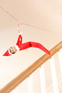This image shows an elf on the shelf doll hanging from a candy cane across a rope like a zipline. This is one of the many elf on the shelf ideas for home included in this post. Look at how much fun this Elf on the Shelf is having by hanging by a candy cane on a zipline. This is one of the 50+ elf on the shelf ideas for home included in this round up. Plus, you’ll get a set of free Elf on the Shelf planning printables. This includes a blank elf on the shelf calendar, list of elf on the shelf ideas, printable elf on the shelf notes, a fillable elf on the shelf note set, and more.