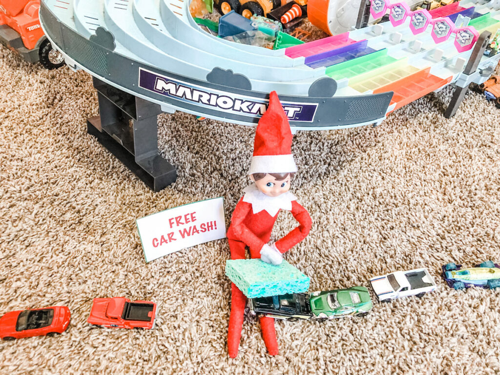 This image shows an elf pretending to wash a toy car. It has a free note sitting next to it that says free car wash! This is one of the many elf on the shelf ideas for home included in this post.