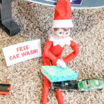 This image shows an elf pretending to wash a toy car. It has a free note sitting next to it that says free car wash! This is one of the many elf on the shelf ideas for home included in this post.